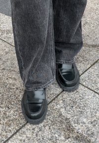 Czarne skórzane loafersy z zaokrąglonym noskiem, przeszyciami i płaską gumową podeszwą, w zestawieniu z szarymi dżinsami na kamiennej powierzchni.