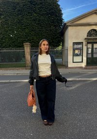Veste en cuir noir, chemise blanche, jean large bleu foncé, sac tissé orange avec un charme en fourrure, et sandales noires. Cadre décontracté en extérieur.