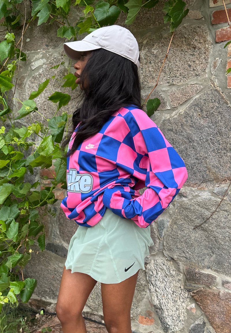 Femme portant une casquette blanche, une chemise Nike à manches longues à carreaux roses et bleus, et un short Nike vert clair, se tient de profil près d’un mur en pierre avec des vignes vertes.