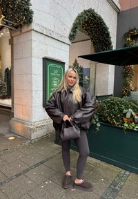 Brązowa oversizeowa kurtka puchowa, czarne legginsy, brązowe buty wyłożone futerkiem, trzyma małą torebkę; miejska sceneria z zielenią i świąteczną dekoracją.