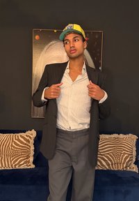 Νεαρό άτομο που φορά ριγέ σακάκι, λευκό πουκάμισο, γκρι παντελόνι και κίτρινο καπέλο, στέκεται μπροστά σε έναν σκοτεινό τοίχο με αφηρημένη τέχνη και μοτίβο μαξιλάρια.