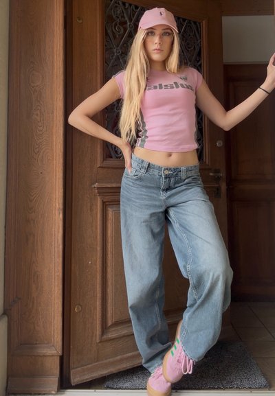 Top corto rosa con texto gráfico, jeans sueltos de ajuste ligero en azul claro y zapatillas rosa con rayas verdes, en un fondo de puerta de madera.