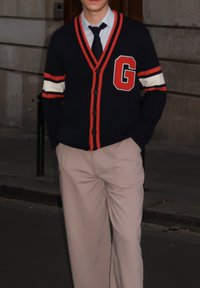Marineblå cardigan med røde detaljer og et stort "G"-emblem, knappet igjen over en lyseblå skjorte og kombinert med beige bukser.