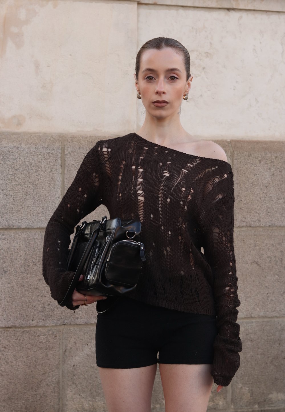 Ciemnobrązowy, dzianinowy sweter o luźnym kroju i postarzanym wyglądzie; czarne szorty; trzymając małą czarną torbę z srebrnymi detalami.