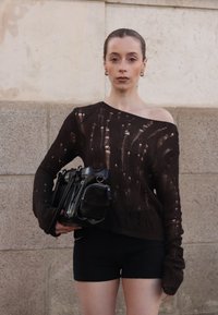 Ciemnobrązowy, dzianinowy sweter o luźnym kroju i postarzanym wyglądzie; czarne szorty; trzymając małą czarną torbę z srebrnymi detalami.
