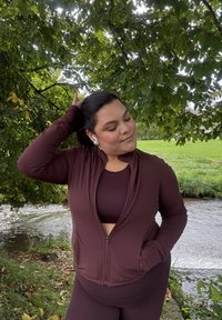 Burgundy sports outfit comprising a zip-up jacket and matching crop top. The fabric looks smooth and fitted, set against a backdrop of lush green foliage.
