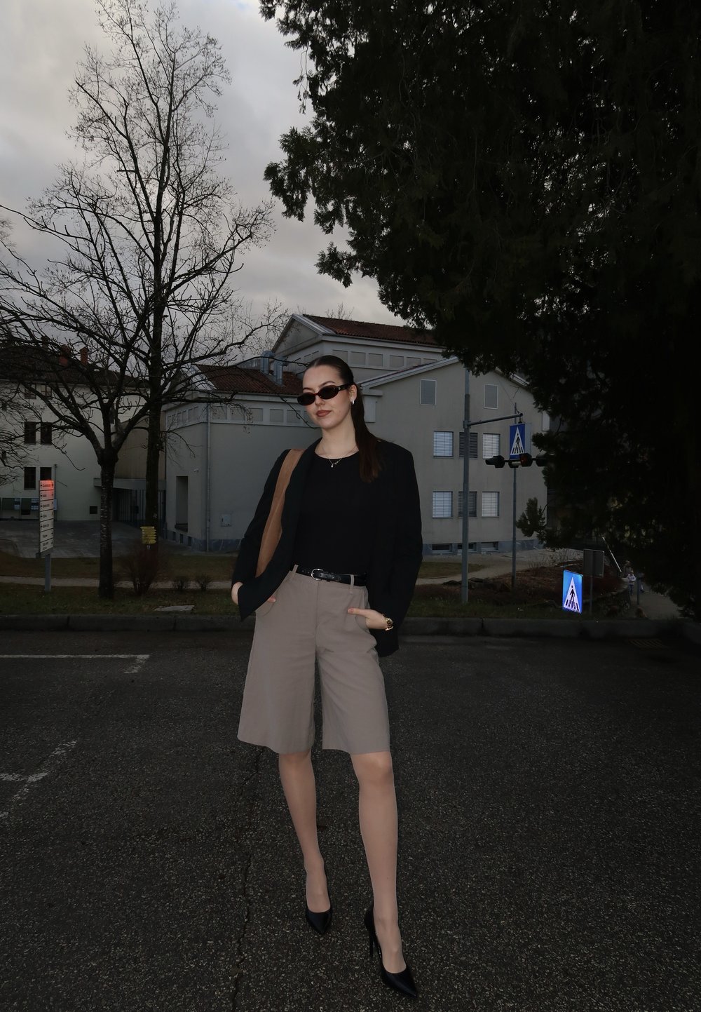 Jeune femme portant des lunettes de soleil, un blazer noir, un short beige et des talons, debout sur une rue pavée avec des arbres sans feuilles et des bâtiments en arrière-plan.