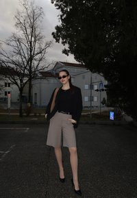 Jeune femme portant des lunettes de soleil, un blazer noir, un short beige et des talons, debout sur une rue pavée avec des arbres sans feuilles et des bâtiments en arrière-plan.