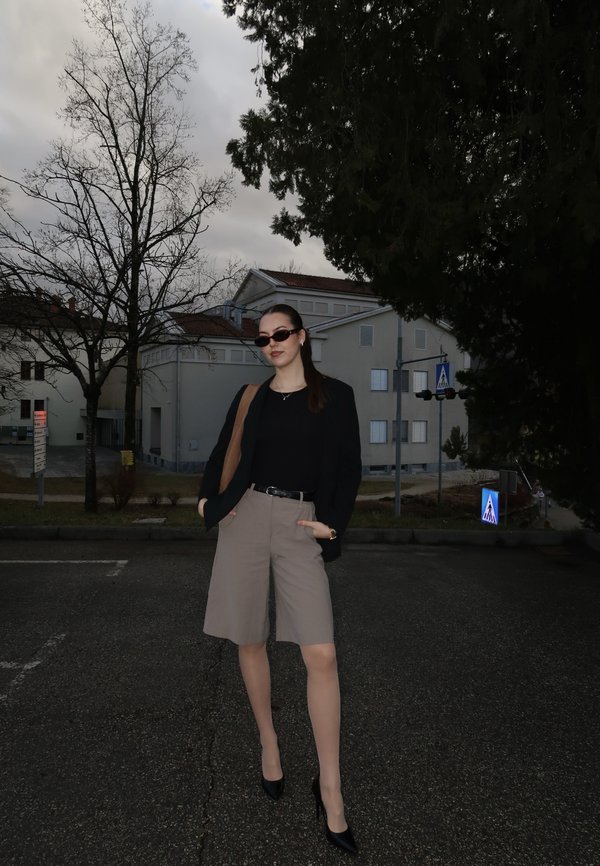 Young woman wearing sunglasses, a black blazer, beige shorts, and heels, standing on a paved street with leafless trees and buildings in the background.
