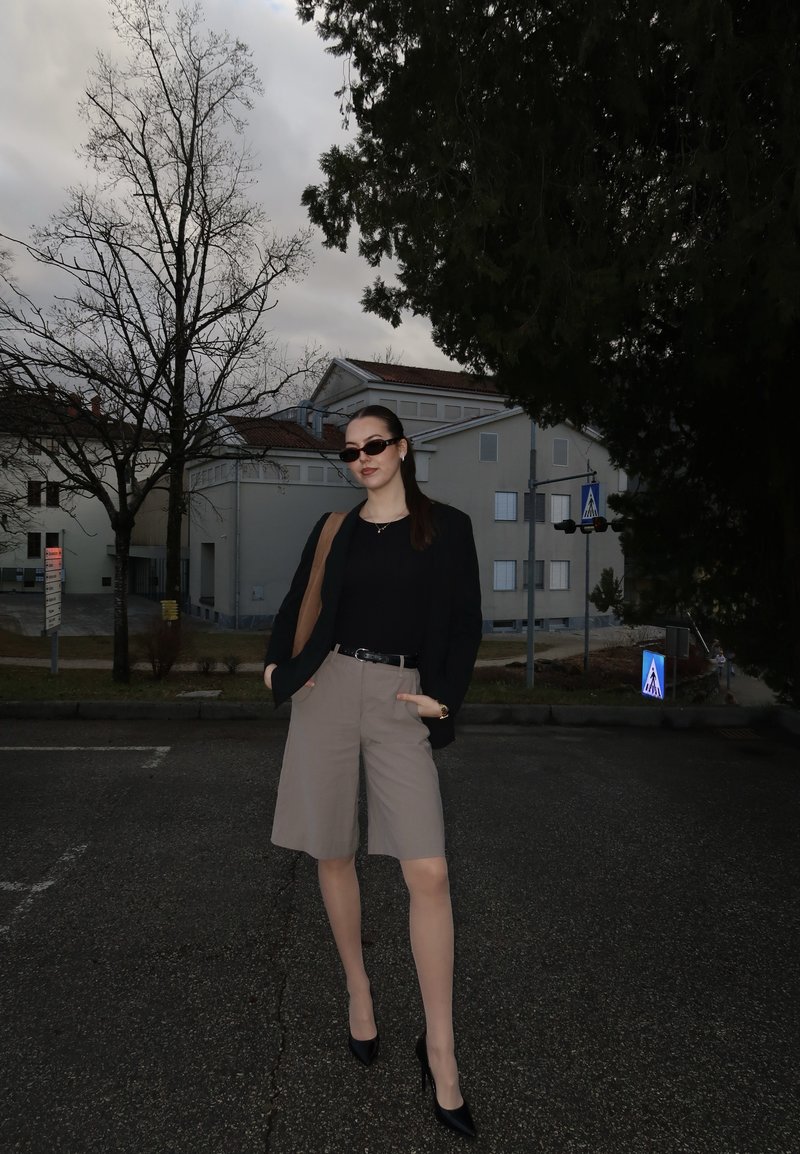 Jeune femme portant des lunettes de soleil, un blazer noir, un short beige et des talons, debout sur une rue pavée avec des arbres sans feuilles et des bâtiments en arrière-plan.