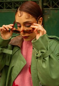 Femme aux cheveux de bébé coiffés ajuste ses lunettes de soleil écaille, vêtue d'un pull rose et d'un manteau en cuir vert avec des détails à boucle.