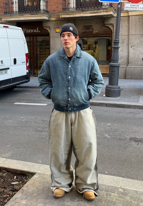 Casaco bomber de denim em azul desbotado, calças de atoalhado cinzentas oversized e botas castanhas. Gorro preto com logótipo branco. Ambiente urbano.