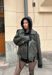 Vihreä nahkainen moottoripyörätakki, jossa on hopeiset yksityiskohdat, epäsymmetrinen vetoketju ja nappisuljenta. Puettu mustan hupparin ja istuvien shortsien päälle.