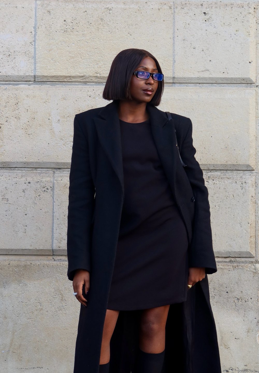 Schwarzer langer Mantel über einem schwarzen Kleid, mit knielangen schwarzen Socken. Die Person hat glattes, dunkelbraunes Haar und trägt eine blaue Brille.
