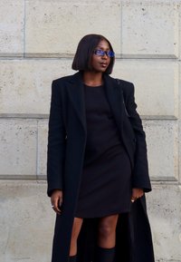 Schwarzer langer Mantel über einem schwarzen Kleid, mit knielangen schwarzen Socken. Die Person hat glattes, dunkelbraunes Haar und trägt eine blaue Brille.