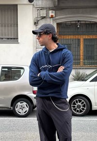 Ναυτικά μπλε φούτερ με λευκό γραφικό, μαύρο παντελόνι cargo με αλυσιδωτή λεπτομέρεια και μαύρο καπέλο. Στηρίζεται δίπλα σε ένα ασημένιο αυτοκίνητο, με τα χέρια σταυρωμένα.