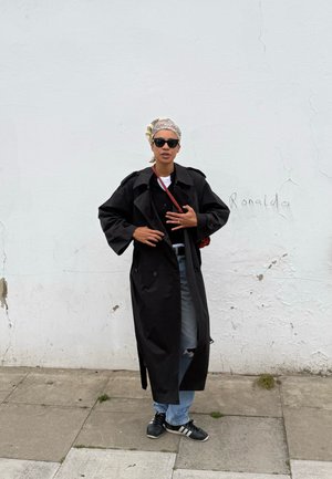 Persona que lleva un abrigo tipo gabardina negro, gafas de sol, pañuelo en la cabeza, vaqueros rotos y zapatillas, de pie en una acera frente a una pared blanca con graffiti.
