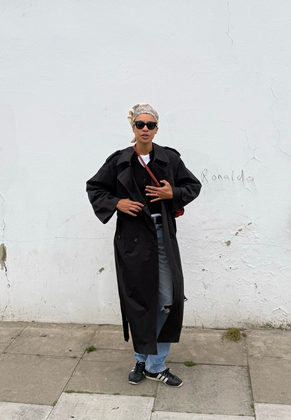Personne portant un trench-coat noir, des lunettes de soleil, un foulard, un jean déchiré et des baskets, debout sur un trottoir contre un mur blanc avec des graffitis.