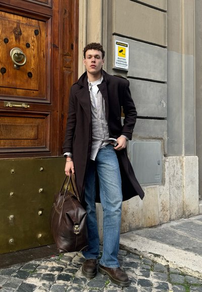 Abrigo de lana negro, camisa blanca de botones, jeans azules, zapatos de cuero marrón y un gran bolso de cuero marrón, frente a una puerta de madera.
