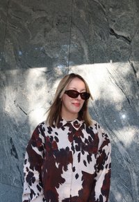 Žena s svijetlosmeškom kosom koja nosi tamne naočale i košulju s uzorkom u smešnim, crnim i bijelim bojama, naslonjena na teksturiranu sivu kamenitu zid.