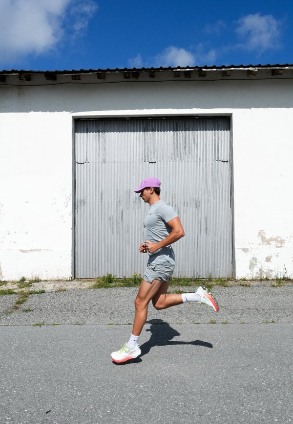 Uomo con cappellino viola e abbigliamento sportivo grigio che corre su una strada lastricata davanti a un edificio bianco con una porta grigia chiusa.
