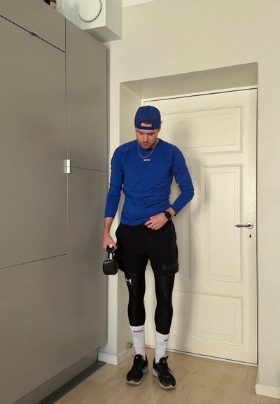 Hombre con camisa azul de manga larga y gorra sosteniendo una pesa rusa, de pie en el interior cerca de una puerta y un armario.