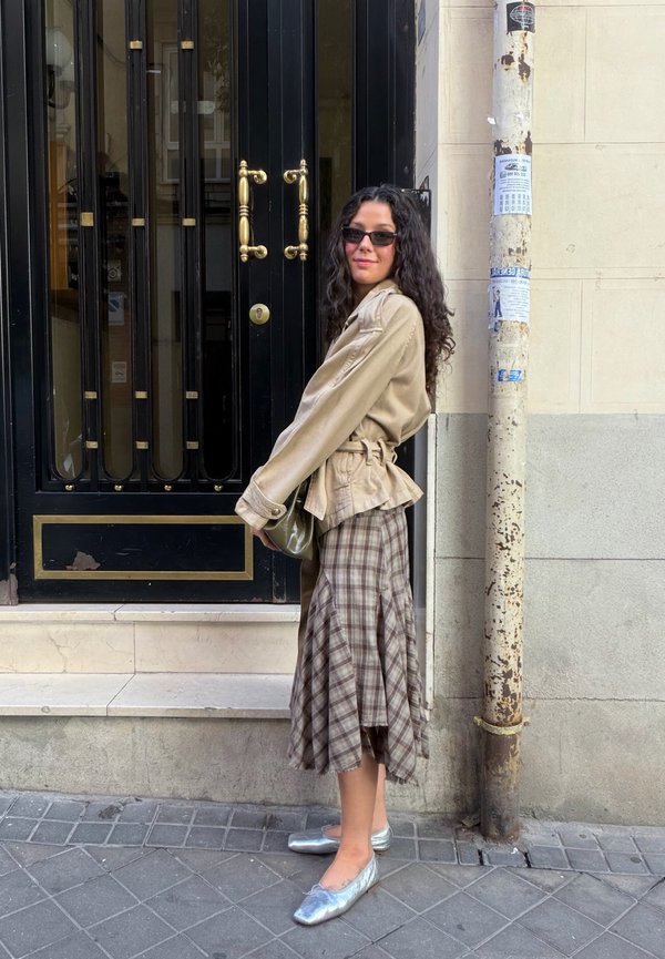 Una mujer con gafas de sol, una chaqueta beige, una falda de cuadros y zapatos planos plateados posa junto a una puerta negra en una acera urbana.