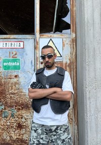Chaleco de cuero negro sobre una camiseta blanca, pantalones cortos de camuflaje y guantes negros. La persona está de pie contra una puerta de metal oxidado con señales de advertencia.