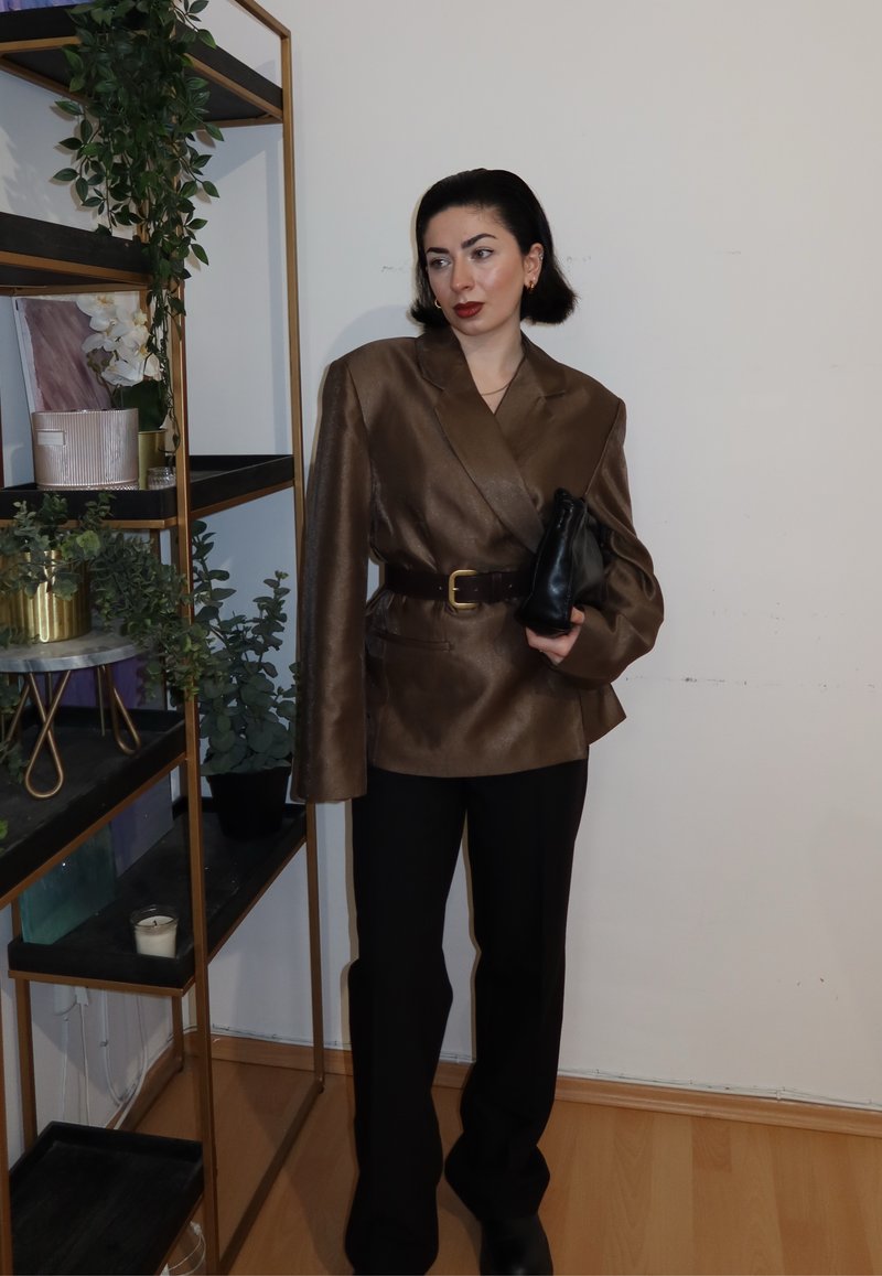 Femme en blazer marron surdimensionné avec ceinture, pantalon noir, tenant une pochette noire, debout à côté d'une étagère dorée avec des plantes et des bougies.