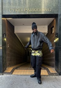 Mustaa nahkaa oleva bomber-takki, jossa on korkea kaulus, keltaiset camouflage-housut ja mustat taistelu-saappaat. Sisäänkäynti merkitty "CYKELPARKERING."