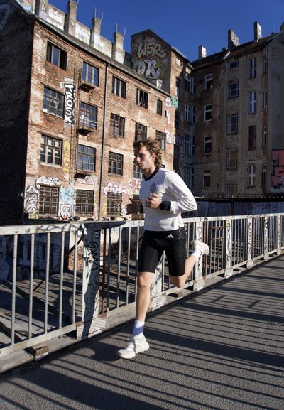 Joven con camisa blanca de manga larga y pantalones cortos negros corriendo en un puente urbano cubierto de grafitis con edificios antiguos de ladrillo al fondo.