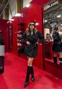 Robe blazer noire à silhouette cintrée, associée à des bottes noires à talons montant jusqu'aux genoux. En arrière-plan, un mur rouge avec des étagères et des miroirs.