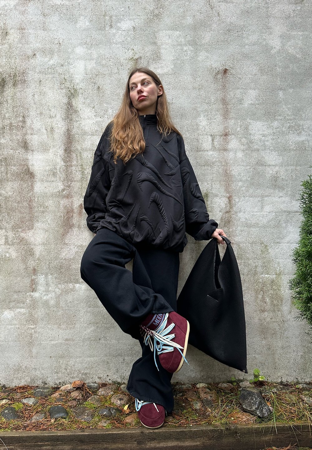 Svart oversized genser med teksturert design, vide svarte bukser og burgunderrøde sko med blå lisser; holder en svart handlenett.