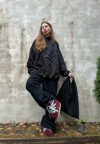 Svart oversized genser med teksturert design, vide svarte bukser og burgunderrøde sko med blå lisser; holder en svart handlenett.