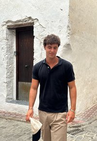 Cămașă polo neagră, pantaloni bej, ținând o pălărie albă. Fundalul prezintă un perete alb texturat și o ușă din lemn cu un mâner metalic.