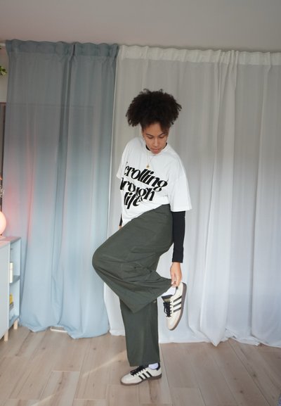 Camiseta blanca con texto negro, pantalones verde oliva y zapatillas beige con rayas negras. Cabello rizado y postura de pie frente al fondo.