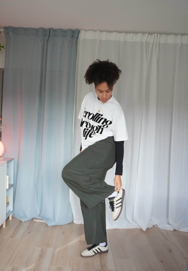 Camiseta branca com texto preto, calças verde-oliva e tênis bege com riscas pretas. Cabelo encaracolado e postura em pé contra um fundo.