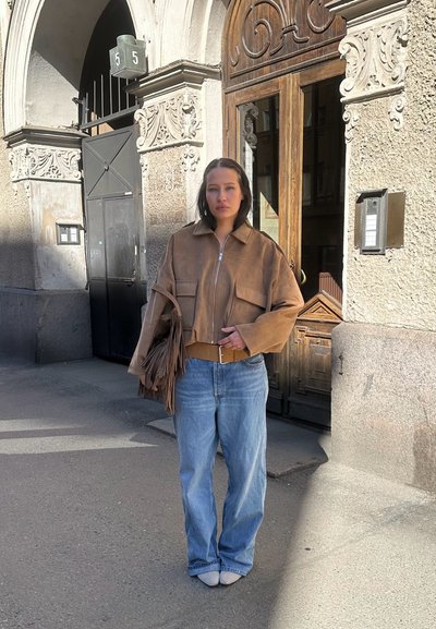 Mujer con chaqueta marrón y vaqueros azules está de pie sosteniendo un bolso con flecos frente a una puerta ornamentada de un edificio con el número 5.