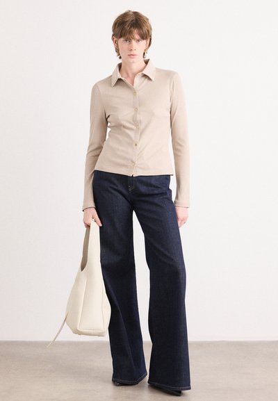 Camisa de manga larga a rayas beige con cuello puntiagudo y botones dorados, combinada con jeans acampanados de un azul oscuro. El modelo sostiene una bolsa de mano blanca.