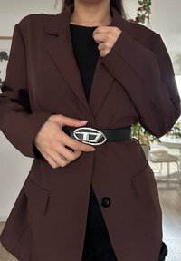Blazer marron à coupe ajustée, top noir en dessous, cintré à la taille avec une ceinture noire brillante ornée d'une boucle ovale argentée.