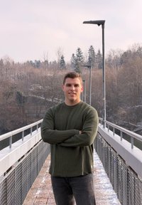 Moški v zelenem rebrastem puloverju, ki samozavestno stoji na sprehajališču z ograjo iz kovine in drevesi v ozadju pod oblačnim nebom.