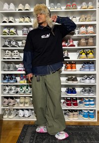 Homme portant un chapeau en fourrure, un T-shirt noir, un pantalon vert et des baskets roses, debout devant des étagères blanches remplies de différentes baskets.