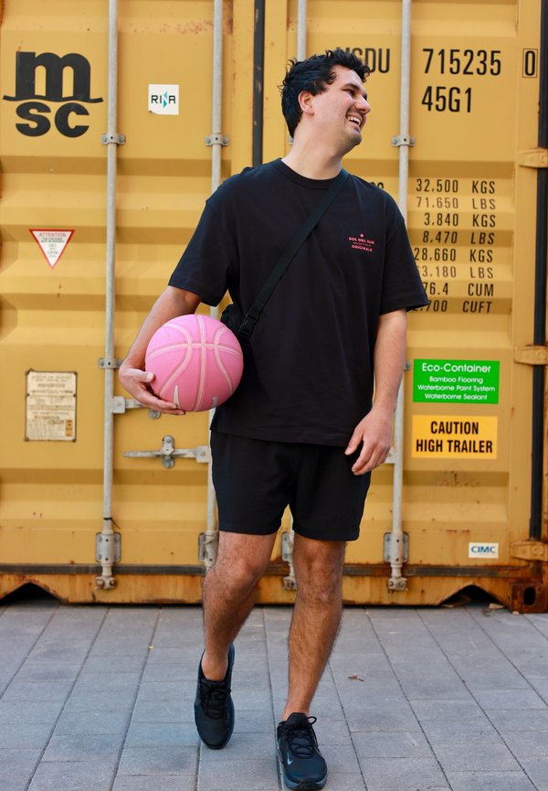 Mand i sort tøj, der holder en pink basketball, smilende og gående foran en gul shippingcontainer med advarselsmærker.