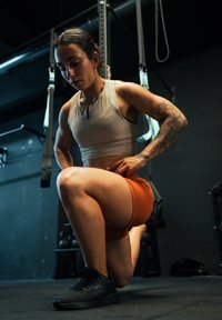Athlète en débardeur beige ajusté et shorts oranges, à genoux sur le sol d'une salle de gym. Chaussures de sport noires et tatouages visibles sur son bras.