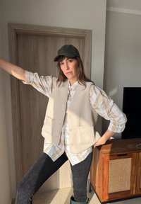 Γυναίκα που φοράει μπλούζα σε μπεζ καρό σχέδιο, κρεμ φλις γιλέκο, μαύρο παντελόνι και σκουρό πράσινο καπέλο, ποζάροντας σε εσωτερικό χώρο κοντά σε ξύλινη πόρτα και ντουλάπι.