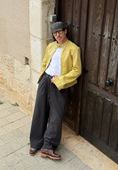 Hombre con chaqueta amarilla, camisa blanca, pantalones anchos negros, zapatos marrones, sombrero gris y gafas apoyado contra una puerta de madera en una acera de piedra.