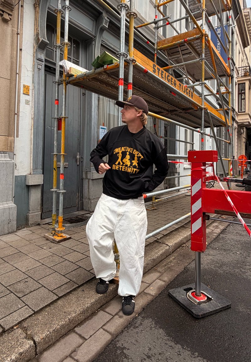 En ung mand i sort kasket, sort sweater og hvide posede bukser står på kantstenen nær orange stilladser og en rød sikkerhedsbarriere på en gade i byen.