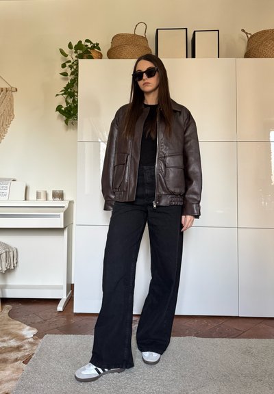 Mujer con chaqueta de cuero marrón oscuro, pantalones anchos negros, zapatillas blancas y gafas de sol negras, de pie en el interior junto a armarios blancos.