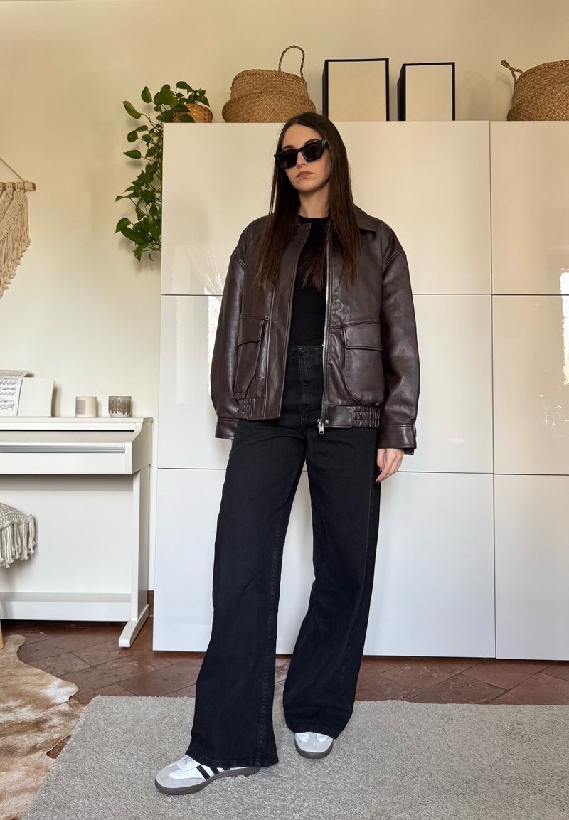 Femme portant une veste en cuir marron foncé, un pantalon noir à jambes larges, des baskets blanches et des lunettes de soleil noires, debout à l'intérieur près de placards blancs.