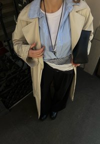 Beženi plašč, svetlo modra srajca preložena čez belo majico s črno-belo črtasto pasom, črne široke hlače in črne čevlje.